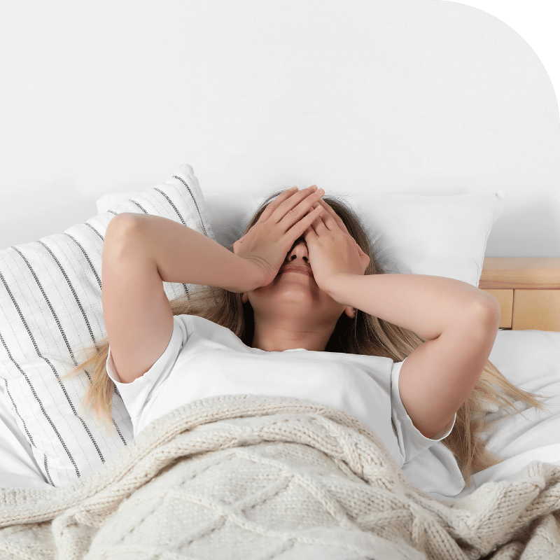 A person with long blonde hair lies in bed, covered with a knitted beige blanket, wearing a white shirt. They have both hands covering their face, resting on a pillow with a white and black striped pillowcase. The background includes a wooden headboard and a white wall with a green section at the top.