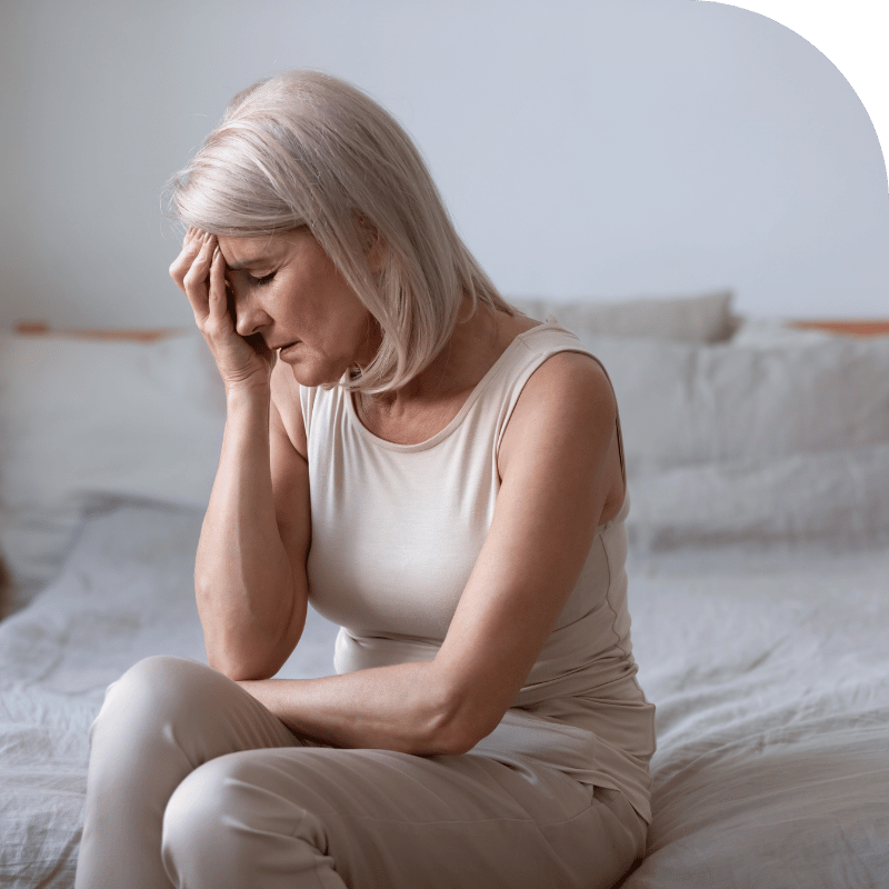 A person with short, light-colored hair sits on a bed, wearing a sleeveless beige top and matching pants. They have one hand on their forehead and the other resting on their lap, with a neutral background featuring a white wall, a green section at the top, and a blurred bed with pillows.