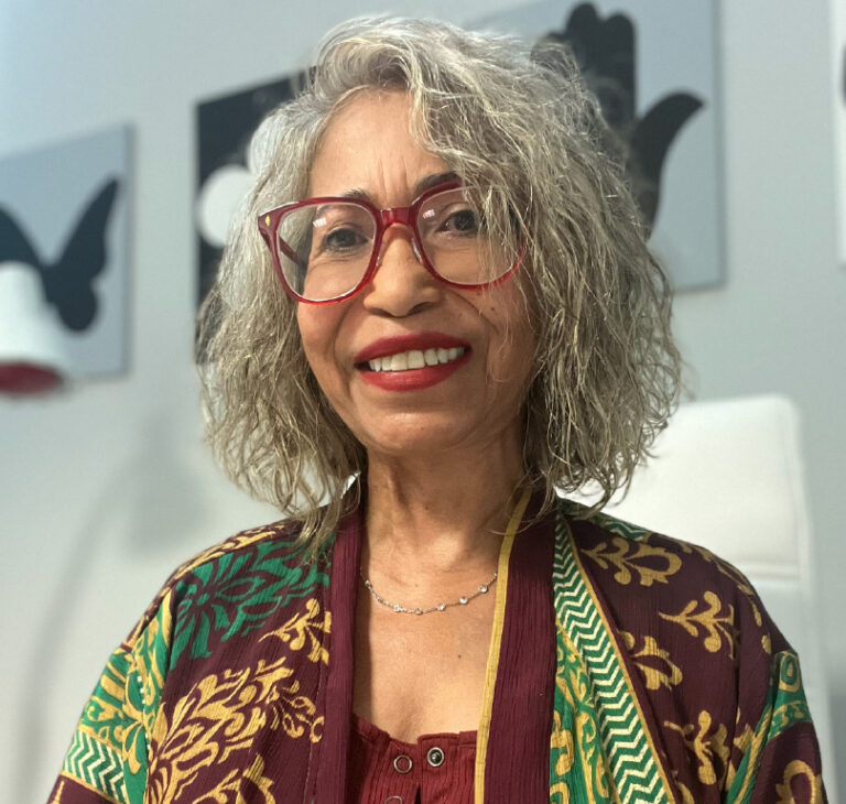 A person with curly gray hair wearing large red-framed glasses and a colorful, patterned robe with green, gold, and maroon designs, smiling warmly. They are seated indoors with a white chair and abstract wall art featuring black shapes and a red lamp in the background.