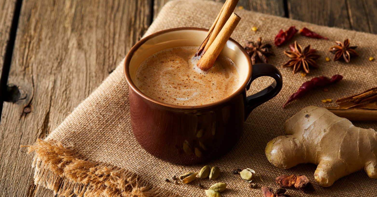 A warm cup of spiced chai on a wooden table, garnished with cinnamon sticks. Surrounding the cup are star anise, dried chili peppers, cardamom pods, and a piece of fresh ginger, all placed on a burlap cloth. The scene evokes a cozy, aromatic atmosphere.