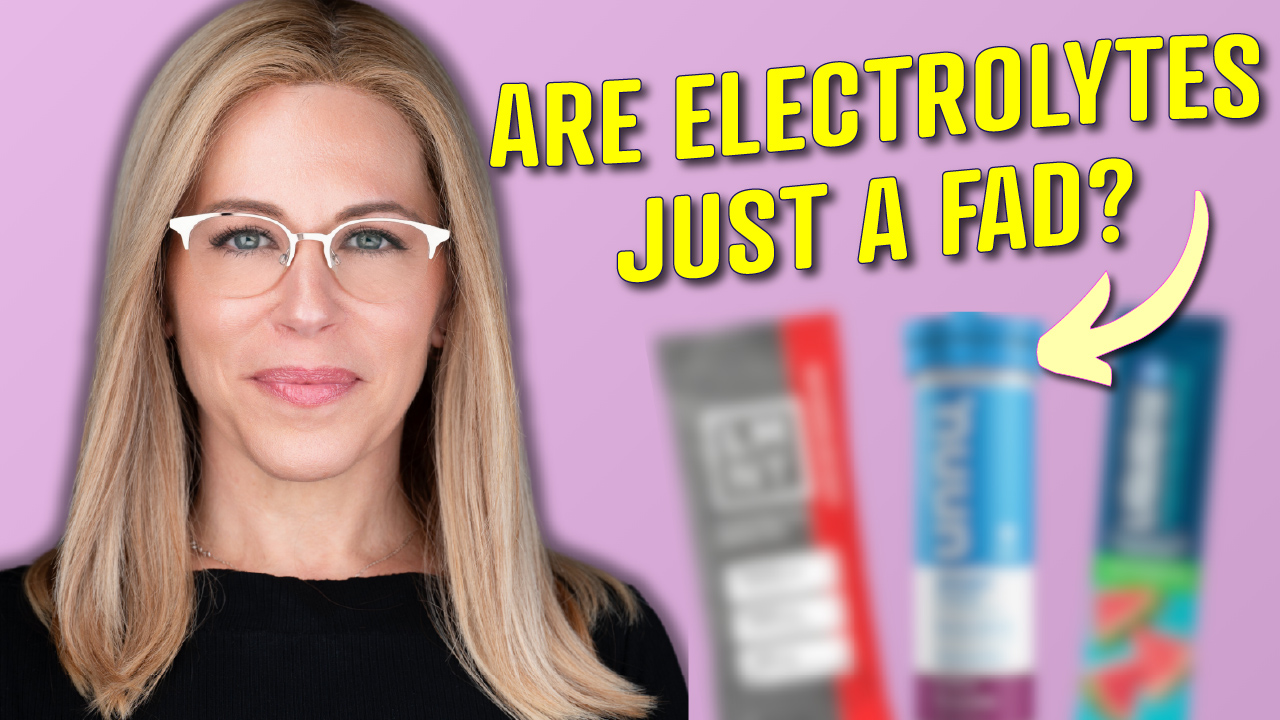 A blonde woman with glasses in a black top is positioned on the left against a pink background . Large yellow text on the right asks, "ARE ELECTROLYTES JUST A FAD?" . Blurred images of three different electrolyte packets/tubes are visible behind the text , and a yellow curved arrow points toward them .